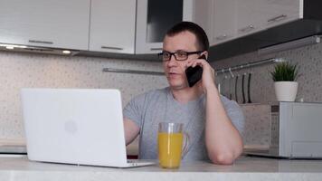 A young man, while working remotely from home, is talking on the phone and looking at a laptop screen while sitting at a table. video