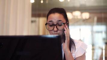 ung skön företag kvinna i glasögon Arbetar använder sig av en mobil dator och mobil telefon Sammanträde på en tabell i de kontor med en Bra humör. närbild. video