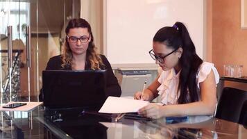 två företag skön företag kvinnor bär glasögon i företag kläder arbetssätt på en företag projekt i ett kontor Sammanträde på en tabell använder sig av en dator. video