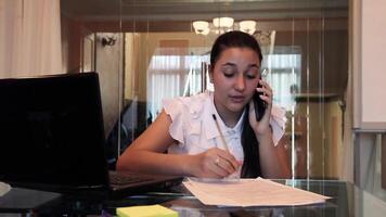 lindo jovem o negócio menina com Sombrio cabelo dentro o negócio roupas falando em uma Móvel telefone discutindo uma o negócio projeto dentro documentos dentro a escritório sentado às uma mesa dentro uma mau humor. hd video