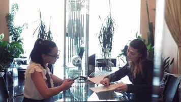 Two business women in business clothes discussing a business project sitting at a table in the office opposite each other using a mobile phone and a computer. video