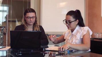 en team av två ung skön företag flickor bär glasögon är arbetssätt på en företag projekt i ett kontor Sammanträde på en tabell använder sig av en mobil dator och dokument. video