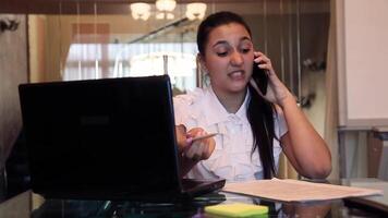 Beautiful young business girl with dark hair in business clothes talking on a mobile phone discussing a business project in an office sitting at a table. HD video