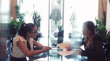 Two business women in business clothes discussing a business project sitting at a table in a beautiful office across from each other using a mobile phone and a computer. HD. video