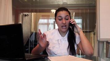 Beautiful young business girl with dark hair in business clothes talking on a mobile phone discussing a business project in an office sitting at a table in a bad mood. HD video