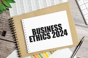 Spiral notebook with BUSINESS ETHICS 2024 text on financial charts. Corporate governance and responsibility concept photo