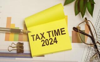 Sticky note with TAX TIME 2024 on financial chart. Taxation and finance reminder concept with pen, glasses, and green plant on office desk photo