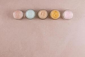 Set of cosmetic cream of various colors in glass jars on beige stone background. Top view. border. A copy space. Layout for product advertising. photo