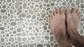 A bird's-eye view of tanned human feet standing on uniquely shaped ash tiles in a bathroom with water splashing and running down the drain. photo