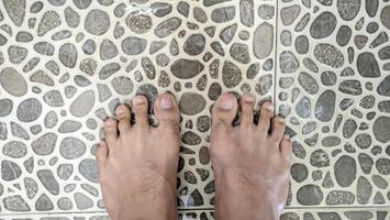 A bird's-eye view of tanned human feet standing on uniquely shaped ash tiles in a bathroom with water splashing and running down the drain. photo