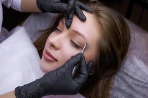 The master uses tweezers to correct the shape of the eyebrows before the permanent makeup procedure. PMU Procedure, Permanent Eyebrow Makeup. photo