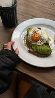 Woman eating sandwich with fried egg and avocado on wooden table in cafe photo