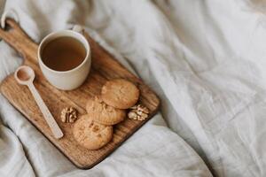 Cup of tea and oatmeal cookies on wooden board on white bedding. photo