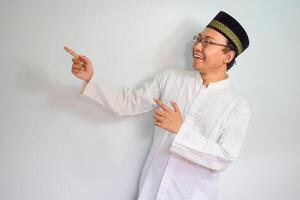 Asian Muslim man wearing glasses and white cloth smiling and pointing pose for ramadhan and eid al fitr. Isolated white background photo