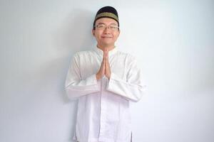 Asian Muslim man wearing glasses and white cloth smiling doing greeting pose for ramadhan and eid al fitr. Isolated white background photo