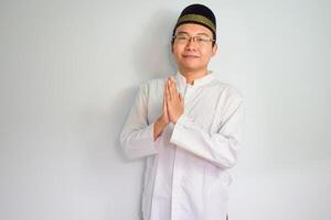 Asian Muslim man wearing glasses and white cloth smiling doing greeting pose for ramadhan and eid al fitr. Isolated white background photo