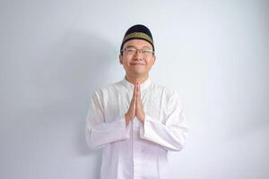 Asian Muslim man wearing glasses and white cloth smiling doing greeting pose for ramadhan and eid al fitr. Isolated white background photo