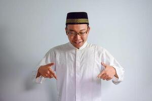 Asian Muslim man wearing glasses and white cloth pointing down pose for ramadhan and eid al fitr. Isolated white background photo