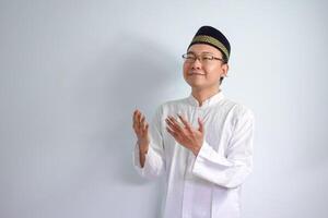 Asian Muslim man wearing glasses and white cloth doing praying  hand pose for ramadhan and eid al fitr. Isolated white background photo