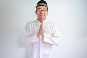 Asian Muslim man wearing glasses and white cloth smiling doing greeting pose for ramadhan and eid al fitr. Isolated white background photo