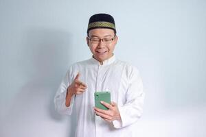 Asian Muslim man wearing glasses and white cloth pointing phone pose for ramadhan and eid al fitr. Isolated white background photo