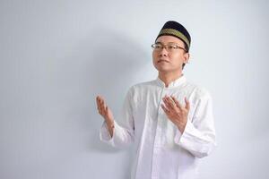 Asian Muslim man wearing glasses and white cloth doing praying  hand pose for ramadhan and eid al fitr. Isolated white background photo