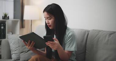 cenas seletivo foco tomada, feliz jovem mulher sentado em sofá usando digital tábua e Móvel telefone para conectados compras dentro vivo quarto às lar, tecnologia dinheiro carteira e conectados Forma de pagamento conceito video