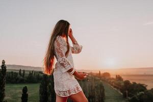 contento mujer en blanco boho vestir en puesta de sol en montañas. romántico mujer con largo pelo en pie con su espalda en el puesta de sol en naturaleza en verano con abierto manos. silueta. naturaleza. puesta de sol. foto