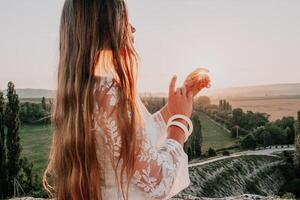 Happy woman in white boho dress on sunset in mountains. Romantic woman with long hair standing with her back on the sunset in nature in summer with open hands. Silhouette. Nature. Sunset. photo