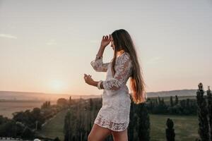 contento mujer en blanco boho vestir en puesta de sol en montañas. romántico mujer con largo pelo en pie con su espalda en el puesta de sol en naturaleza en verano con abierto manos. silueta. naturaleza. puesta de sol. foto