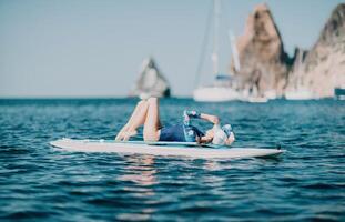 Woman sup yoga. Middle age sporty woman practising yoga pilates on paddle sup surfboard. Female stretching doing workout on sea water. Modern individual female hipster outdoor summer sport activity. photo