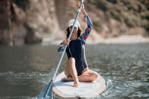 Woman sup yoga. Middle age sporty woman practising yoga pilates on paddle sup surfboard. Female stretching doing workout on sea water. Modern individual female hipster outdoor summer sport activity. photo