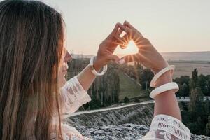 contento mujer en blanco boho vestir haciendo corazón firmar con manos en puesta de sol en montañas. romántico mujer con largo pelo en pie con su espalda en el puesta de sol en naturaleza en verano con abierto manos. foto