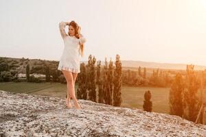 Happy woman in white boho dress on sunset in mountains. Romantic woman with long hair standing with her back on the sunset in nature in summer with open hands. Silhouette. Nature. Sunset. photo