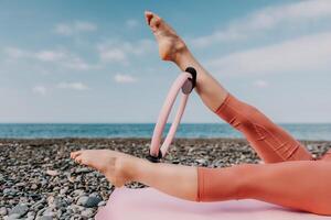 Woman sea pilates. Sporty happy middle aged woman practicing fitness on beach near sea, smiling active female training with ring on yoga mat outside, enjoying healthy lifestyle, harmony and meditation photo