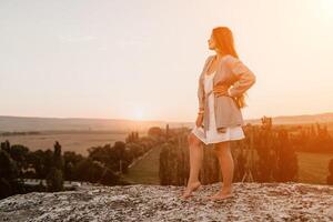 contento mujer en pie con su espalda en el puesta de sol en naturaleza en verano con abierto manos. romántico hermosa novia en blanco boho vestir posando con montañas en puesta de sol foto