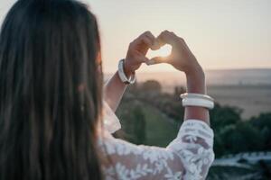 contento mujer en blanco boho vestir haciendo corazón firmar con manos en puesta de sol en montañas. romántico mujer con largo pelo en pie con su espalda en el puesta de sol en naturaleza en verano con abierto manos. foto