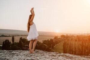 Happy woman standing with her back on the sunset in nature in summer with open hands. Romantic beautiful bride in white boho dress posing with mountains on sunset photo