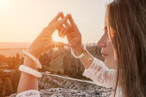 Happy woman in white boho dress making heart sign with hands on sunset in mountains. Romantic woman with long hair standing with her back on the sunset in nature in summer with open hands. photo