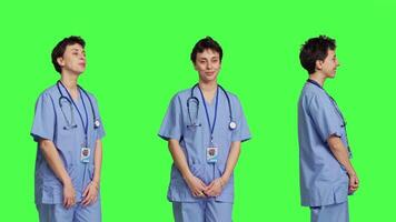 Portrait of medical assistant smiling and posing with confidence, standing against greenscreen backdrop. Nurse wears blue scrubs and stethoscope, feeling successful with health expertise. Camera B. video