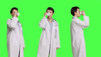 Woman physician drinking a cup of coffee against greenscreen backdrop, taking a break from medical work. Specialist serving caffeine refreshment in studio, wearing a white coat. Camera B. video