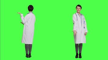 Woman medic does web advertisement against greenscreen backdrop, pointing to left and right sides in studio. Doctor with white coat advertising something for a commercial. Camera A. video