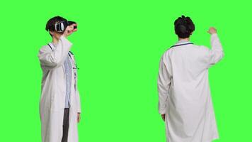 Woman medic works with 3d virtual reality headset in studio, standing against greenscreen backdrop. General practitioner uses vr glasses with interactive vision tool, futuristic exam. Camera B. video