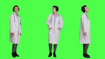 Joyful physician dancing around in the studio with music on headset, having fun with modern cool songs against greenscreen backdrop. Medic woman enjoying audio tunes and singing. Camera A. video