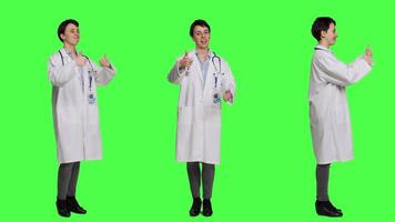 Cheerful medic doing thumbs up symbol against greenscreen backdrop, expresses positivity with like sign. General practitioner with coat giving approval and being satisfied with success. Camera B. video