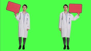 Woman doctor holding a red empty speech bubble sign in studio, showing an isolated blank cardboard icon against greenscreen backdrop. Physician creating announcement with banner. Camera A. video