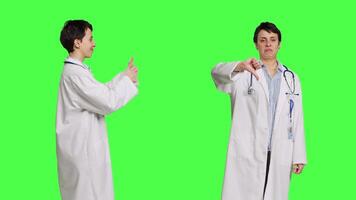 General practitioner showing both like and dislike symbol in studio, expressing approval and disapproval against greenscreen backdrop. Woman medic feels pleased and displeased. Camera B. video