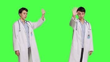 Woman medic waving hello and asking people to come over at checkup, inviting patient to come closer and greets them. Skilled physician with white coat and stethoscope against greenscreen. Camera B. video