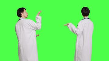 Physician pointing to the left or right to create web commercial standing against greenscreen backdrop, doing a marketing advertisement in studio. Woman medic in white coat shows an ad. Camera B. video