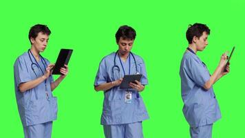 medico assistente navigazione in linea pagine web su tavoletta e sms, utilizzando sociale media applicazioni per Chiacchierare con le persone. infermiera naviga Internet su gadget, in piedi contro schermo verde sfondo. telecamera b. video